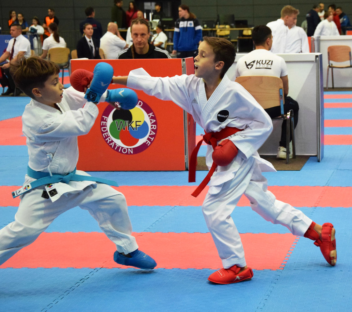 Dwóch zawodników w białych strojach karate walczy ze sobą, chłopiec w czerwonych rękawicach wyprowadza cios, w tle publiczność.