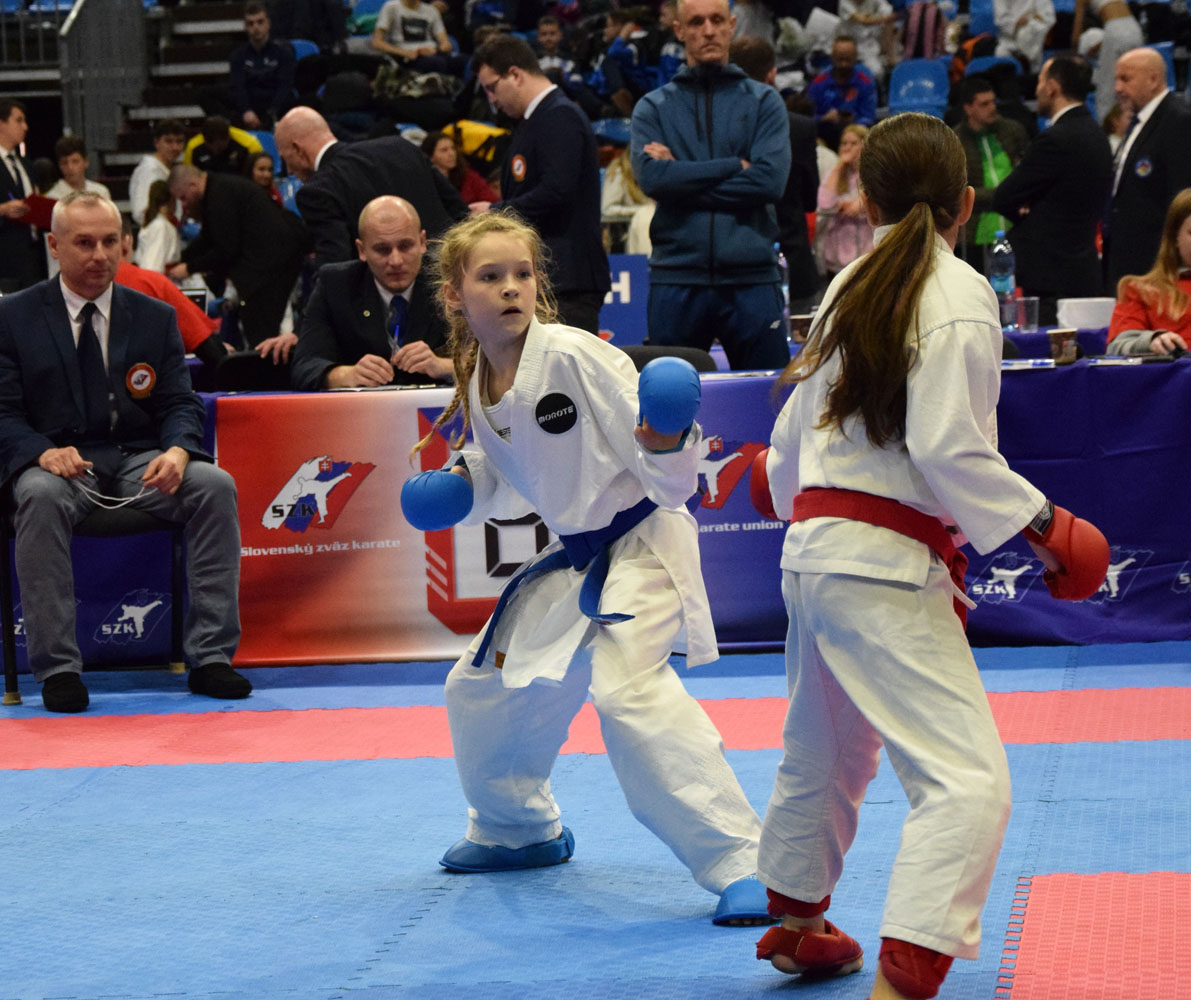 Dwie dziewczynki w strojach karate stoją naprzeciwko siebie na macie, w tle widownia