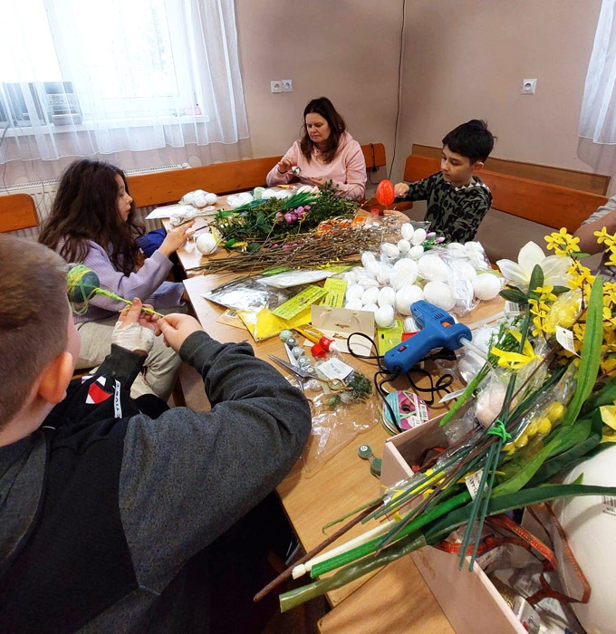Przy stole siedzi trójka dzieci i jedna kobieta, na stole leżą jajka, kwiaty, różne ozdoby