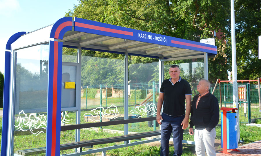 Obok przystanku autobusowego stoi dwóch mężczyzn. 
