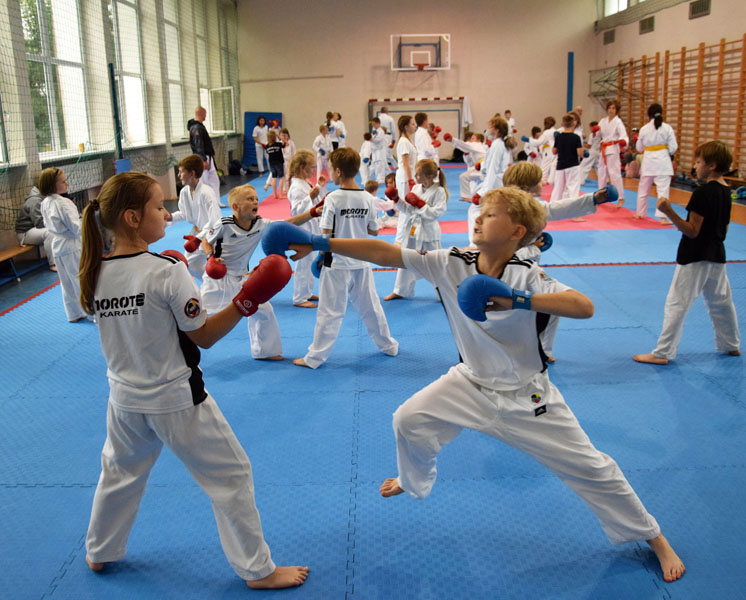 Na sali gimnastycznej grupa młodzieży trenuje karate. Na pierwszym planie chłopiec i dziewczynka walczą.
