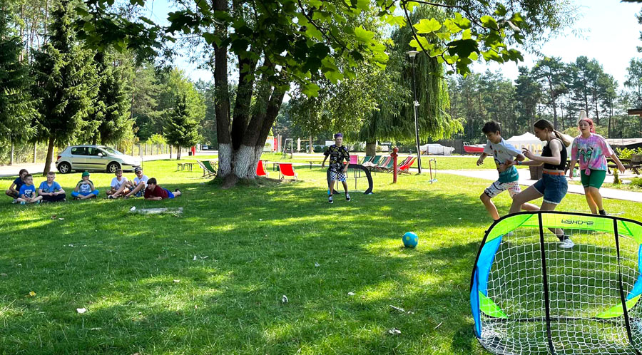 Na trawniku część dzieci siedzi i przygląda się a część gra w piłkę nożną.