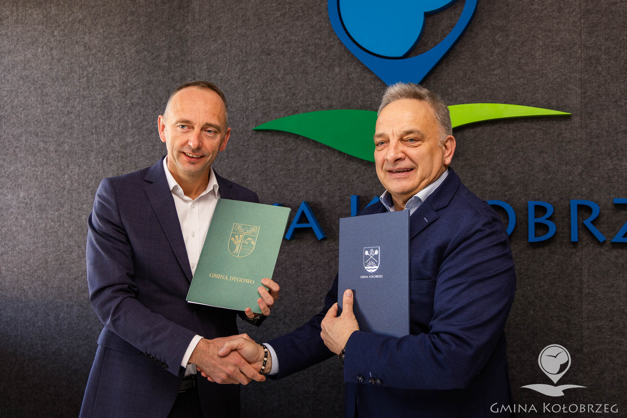 Dwaj mężczyźni podają sobie ręce i prezentują podpisane dokumenty; na okładkach widoczne nazwy gmin. W tle napis &bdquo;Gmina Kołobrzeg&rdquo;, scena przedstawia zawarcie porozumienia między samorządami i oficjalne potwierdzenie wsp&oacute;łpracy.