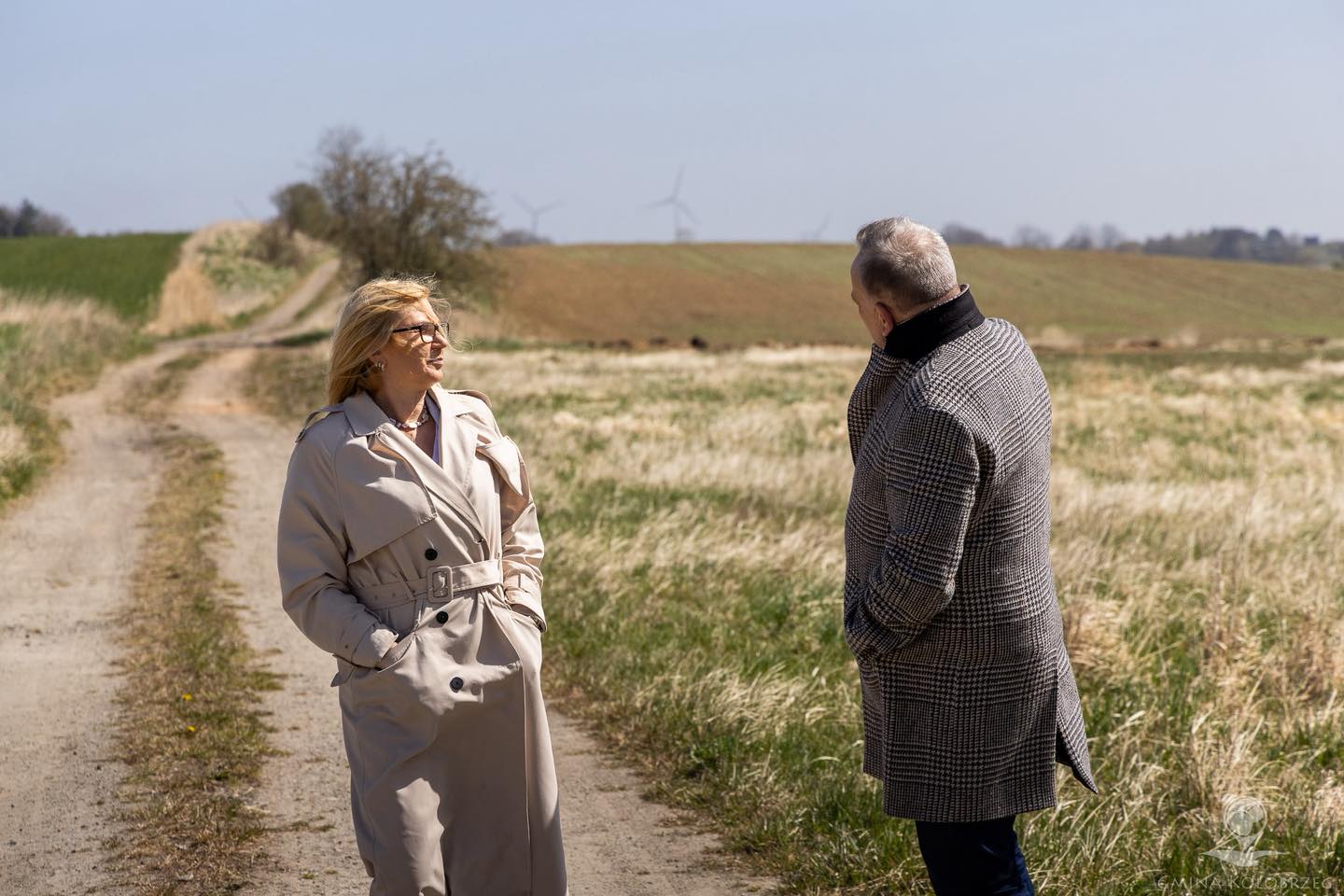 Kobieta i mężczyzna stoją na polnej drodze i rozmawiają, w tle widać pola uprawne oraz turbiny wiatrowe na horyzoncie.