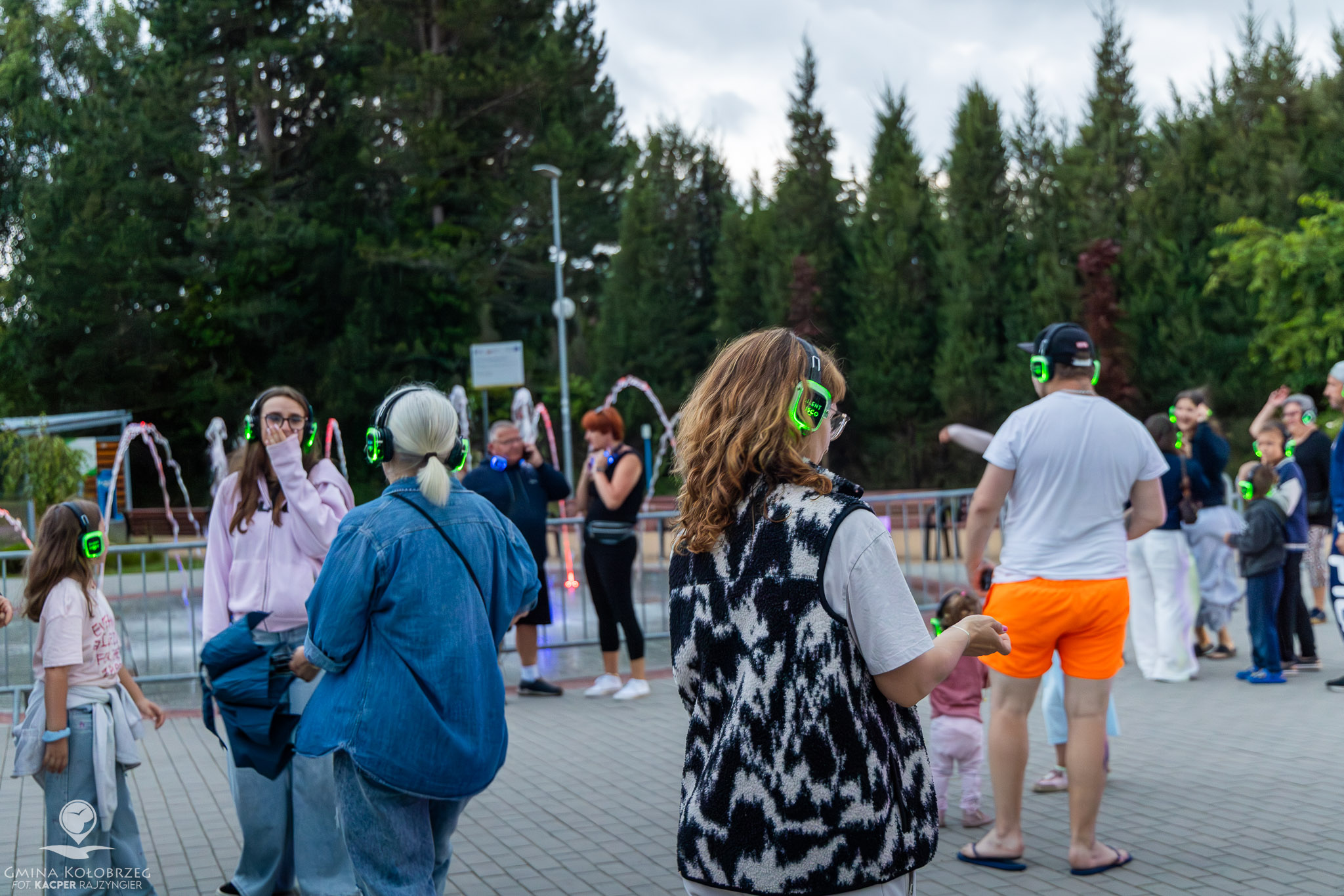 Zdjęcie tańczących ludzi podczas Silent Disco w Grzybowie Ujęcie przedstawia uczestników Silent Disco w słuchawkach, bawiących się na świeżym powietrzu przy podświetlonej fontannie. W tle widoczna jest zieleń oraz grupa osób w różnym wieku korzystających z atrakcji.