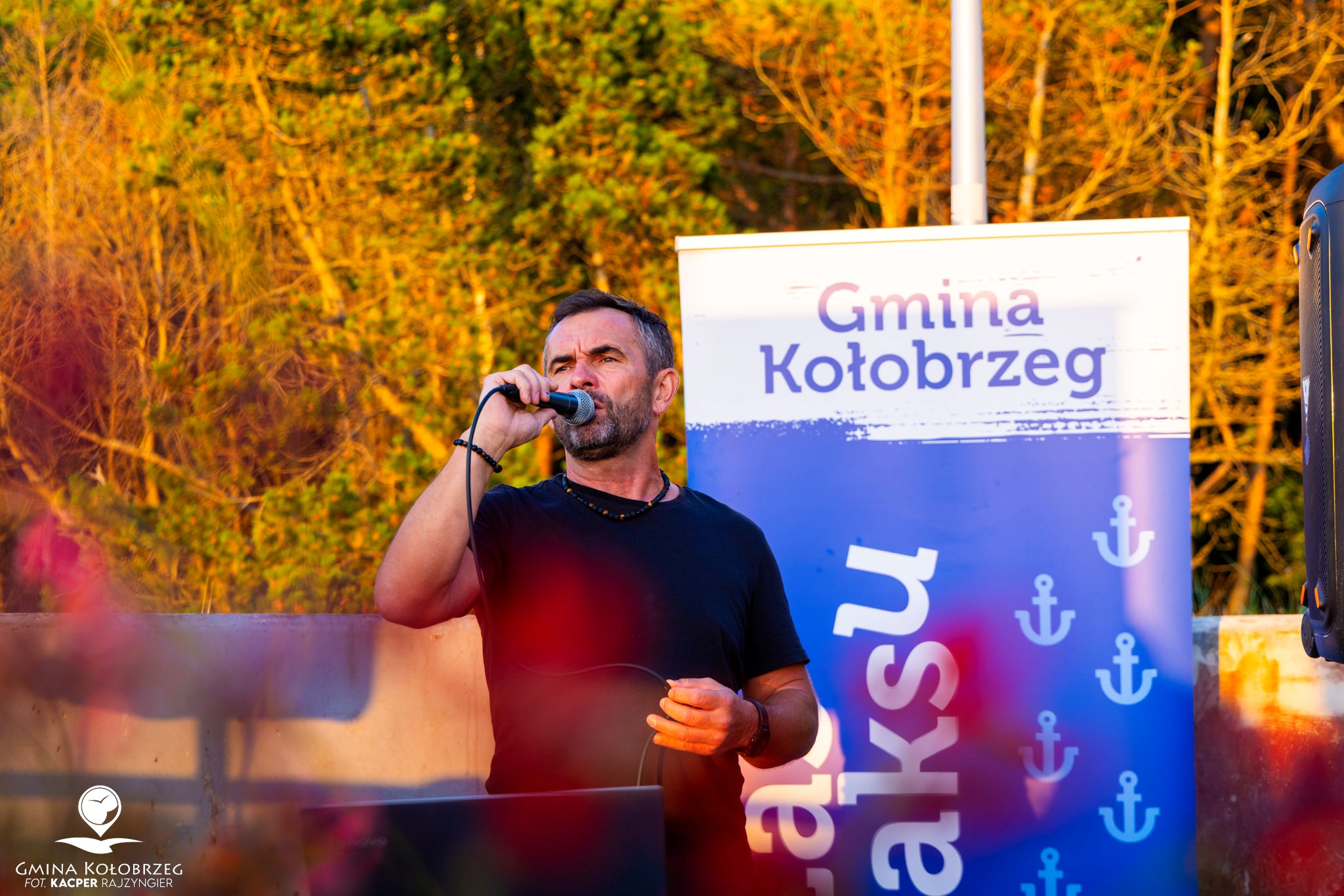 Zdjęcie Krzysztofa Przerady podczas Koncertu Zdjęcie przedstawia osobę prowadzącą wydarzenie plenerowe z mikrofonem. W tle widoczny jest baner „Gmina Kołobrzeg” z motywami wakacyjnymi oraz zieleń oświetlona ciepłym, wieczornym światłem.